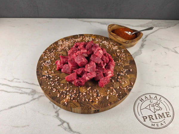 Raw beef chunks on a wooden cutting board with spices, on a marble surface, featuring Halal Prime Meat logo.