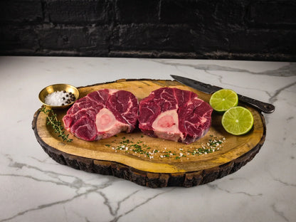 A photograph of two raw beef shanks with prominent marrow bones on a rustic wooden platter accompanied by vibrant green lime halves, a small gold bowl of salt and peppercorns, and a sprig of fresh thyme on a marble surface against a black brick backdrop