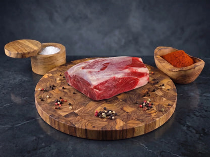 Photograph of a raw marbled beef roast on a round wooden cutting board featuring scattered multicolored peppercorns, a wooden bowl of coarse white salt, and a vessel of vibrant red paprika set against a dark textured background