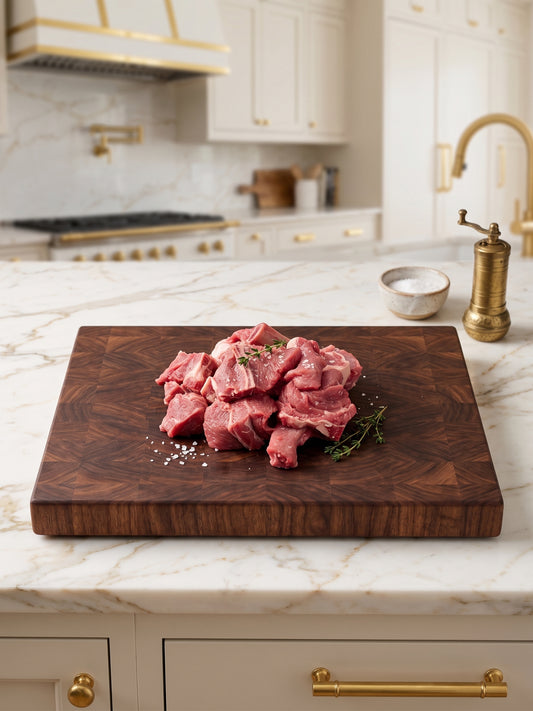 Digital photograph of raw cubed meat on a dark end-grain wooden cutting board in a high-end kitchen with white marble countertops, a brass faucet, coarse salt flakes, and fresh thyme sprigs