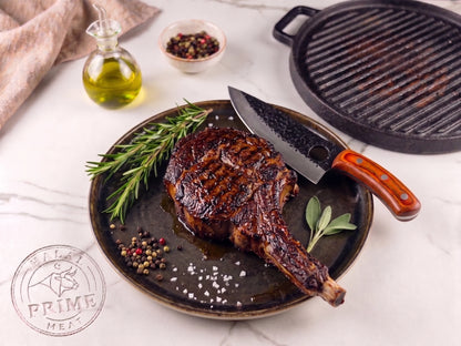 Grilled Cowboy steak on a plate with a knife, rosemary, and spices, next to a grill pan on a white surface.