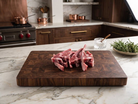 Digital photograph of raw lamb chops on a checkered walnut end-grain cutting board in a luxury kitchen featuring a Wolf stove, copper cookware, and fresh green rosemary sprigs on marble countertops