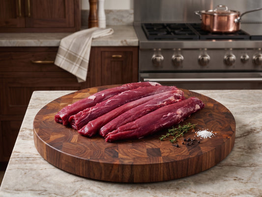 A professional photograph of raw beef tenderloins displayed on a circular walnut end-grain cutting board in a luxury kitchen alongside a stainless steel stove, copper pot, fresh green thyme, and scattered white sea salt crystals
