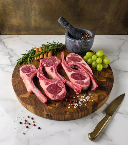 Raw lamb chops on a wooden cutting board with herbs, spices, and a knife on a marble surface.