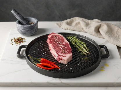 Raw steak on a black grill pan with herbs and peppers on a marble surface.