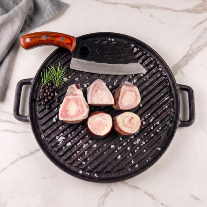 Professional culinary photograph of raw beef marrow bones and a hand-forged cleaver arranged on a black cast iron grill pan over a white marble surface, accented with rosemary sprigs, dark juniper berries, and coarse sea salt crystals