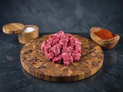 Raw beef stew pieces on a wooden cutting board with salt and pepper bowls on a dark surface