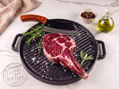 Raw cowboy steak on a grill pan with rosemary, olive oil, and pepper on a marble surface.