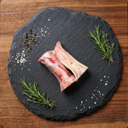 Professional studio photograph of raw beef marrow bones on a black slate tray featuring vibrant green rosemary sprigs and scattered white sea salt crystals against a dark wood background