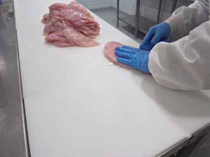A person wearing a white protective coat and blue gloves handles a raw chicken on a white cutting board, with additional sliced raw meat piled nearby in a food processing setting.